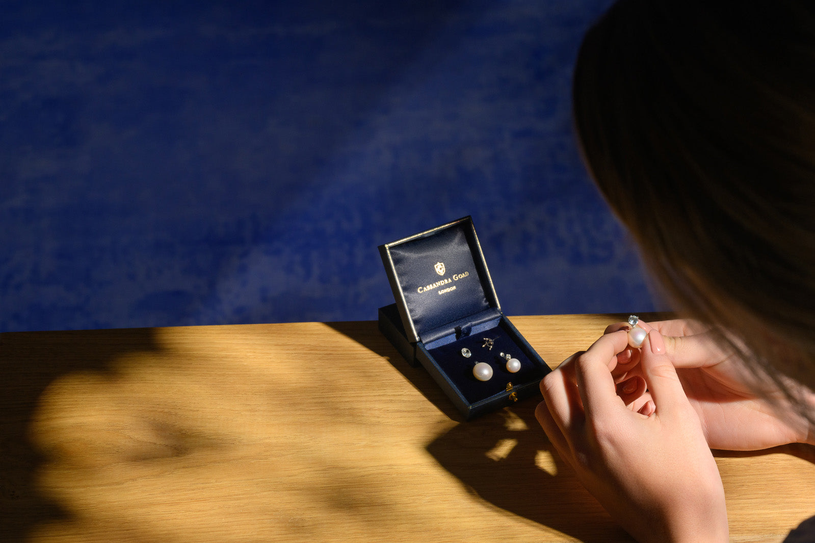 A jewellery box featuring handcrafted pearl earrings with precious stones set in white gold. A person is holding one earring from a matching pair, showcasing it in their hands.