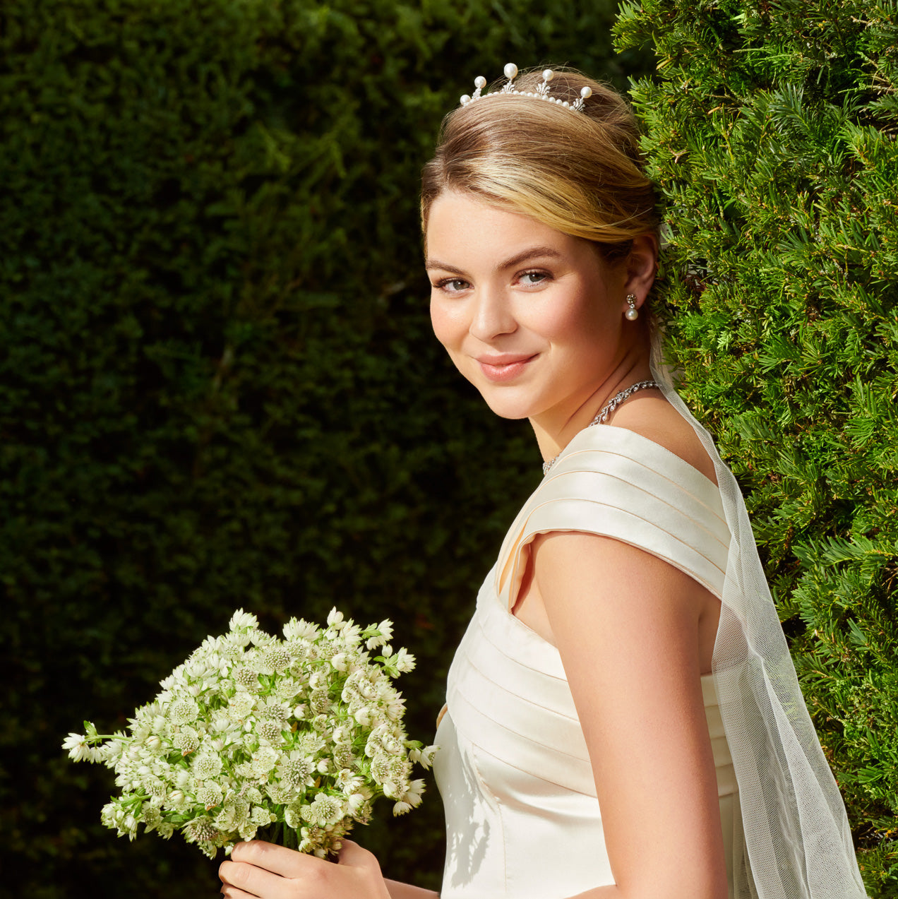 Bride wearing a tiara, earrings and a necklace, all adorned with precious stones and a diamonds, holding a bouquet of white flowers.