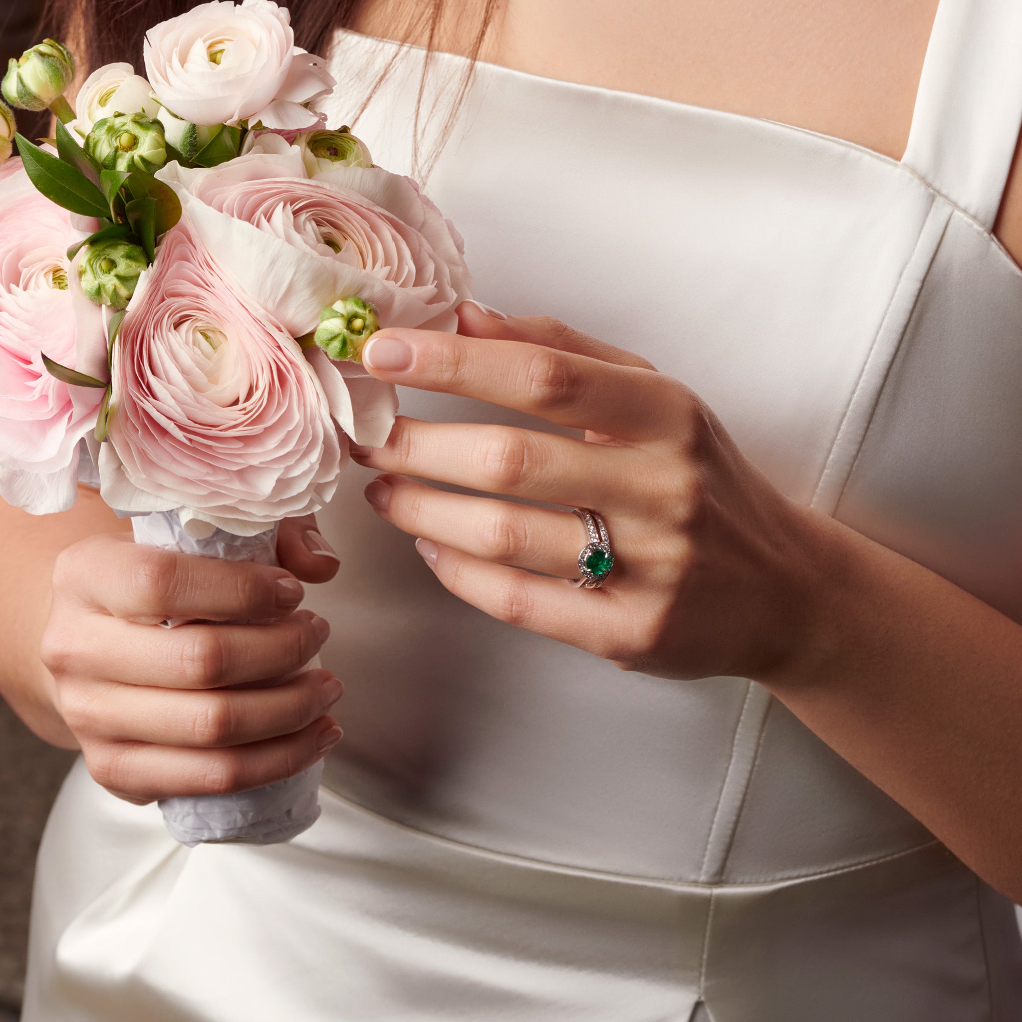 Model wearing a handcrafted 18ct white gold engagement ring with a round  emerald at the centre, surrounded by smaller diamonds. The band of the engagement ring is also set with small diamonds.