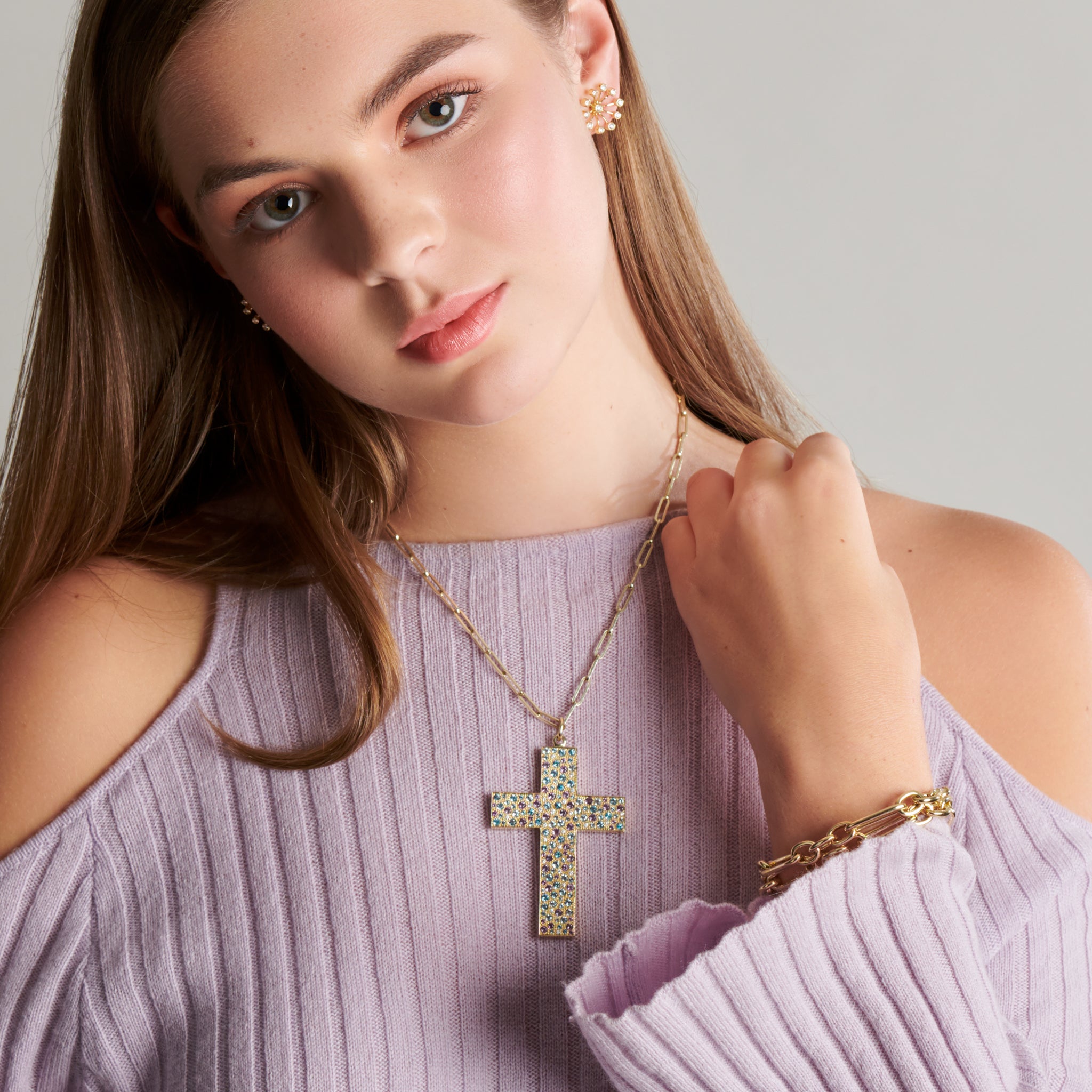 Model wearing a handcrafted yellow gold cross necklace pendant  set with purple and blue precious stones on a gold chain. 