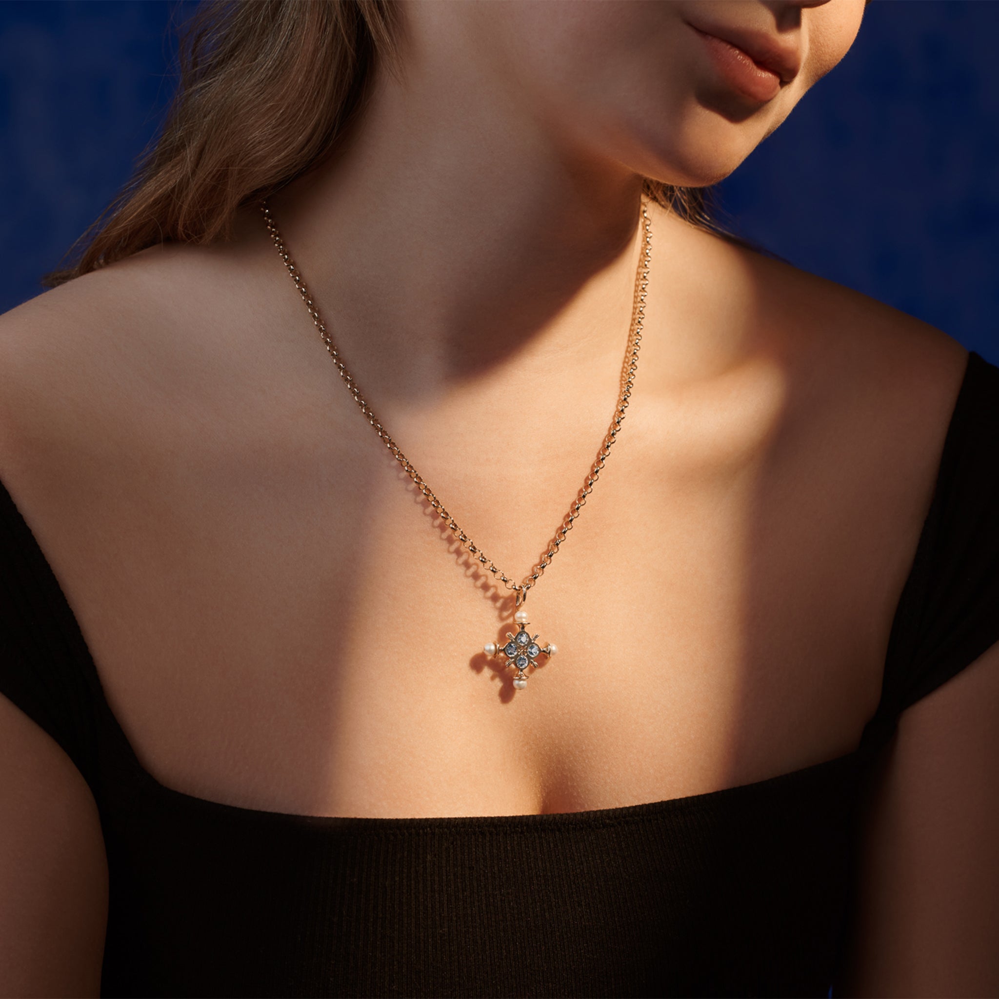 Model wearing a handcrafted cross necklace pendant in yellow gold with a symmetrical arrangement of blue topaz and pearls. 