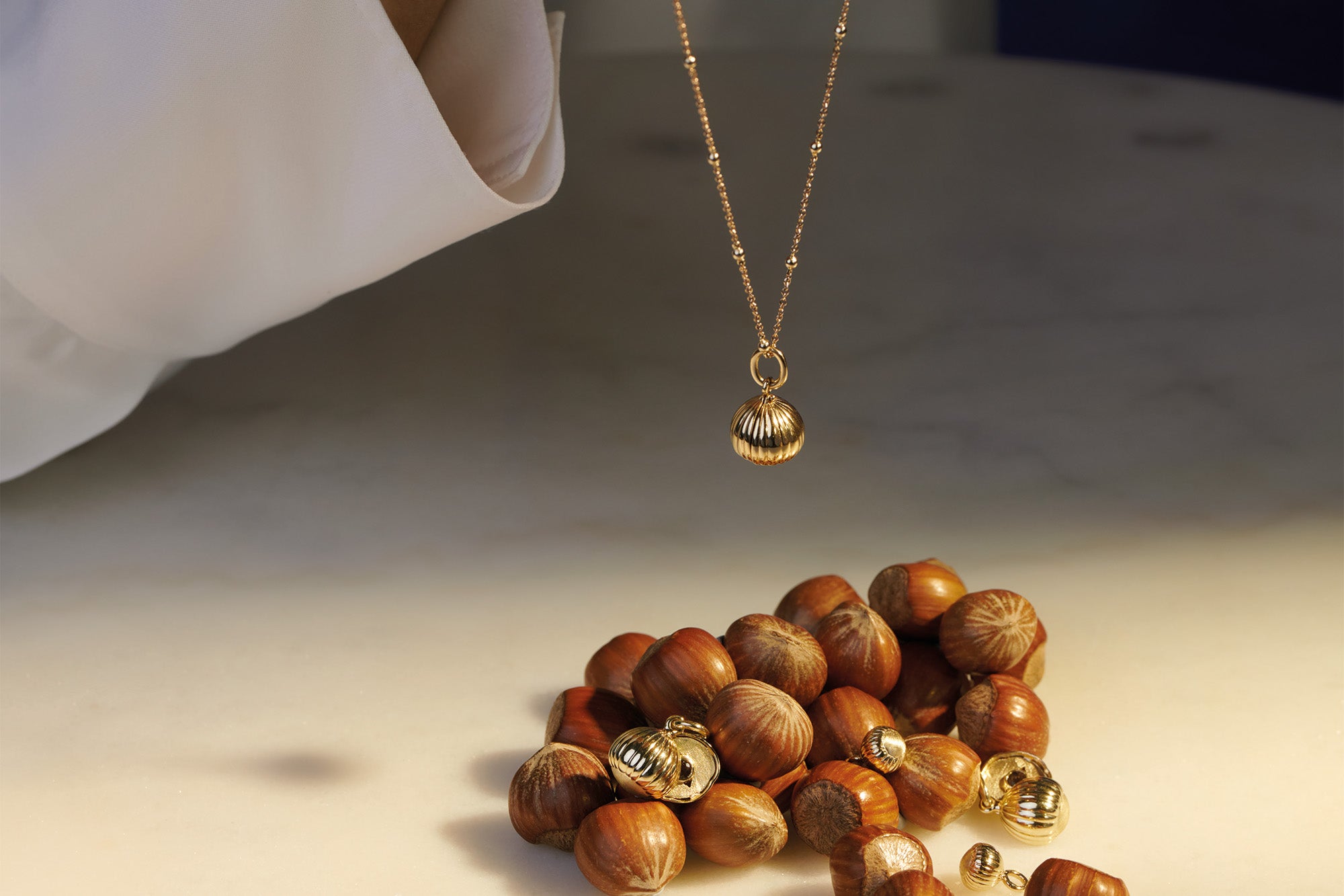 Model holding a yellow gold necklace pendant in the shape of a hazelnut above some hazelnuts. The Noisette design swivels to reveal the centre precious stone.