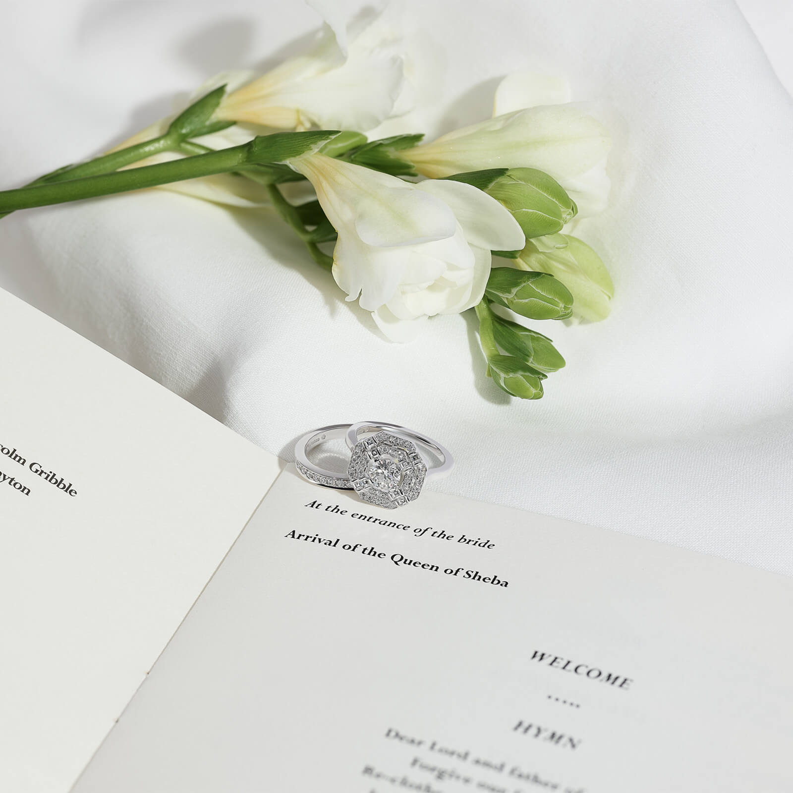 A handcrafted 18ct white gold engagement ring adorned with precious stones, resting on a wedding invite guide next to a flower.