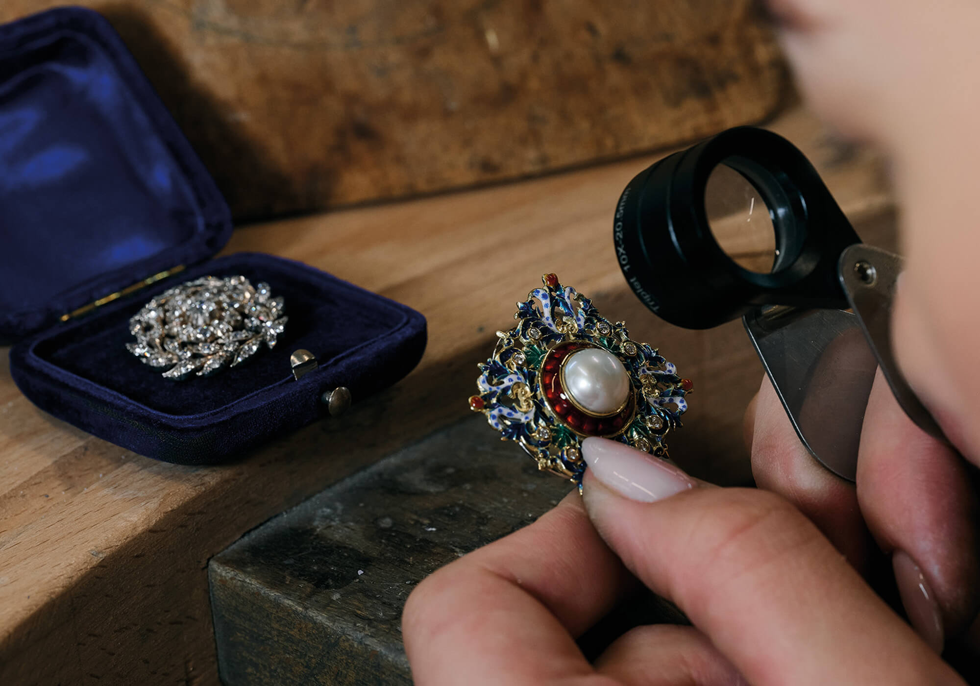 A yellow gold jewellery brooch featuring a central pearl surrounded by blue, red, and green enamel, being valued alongside another yellow gold brooch set with diamonds.