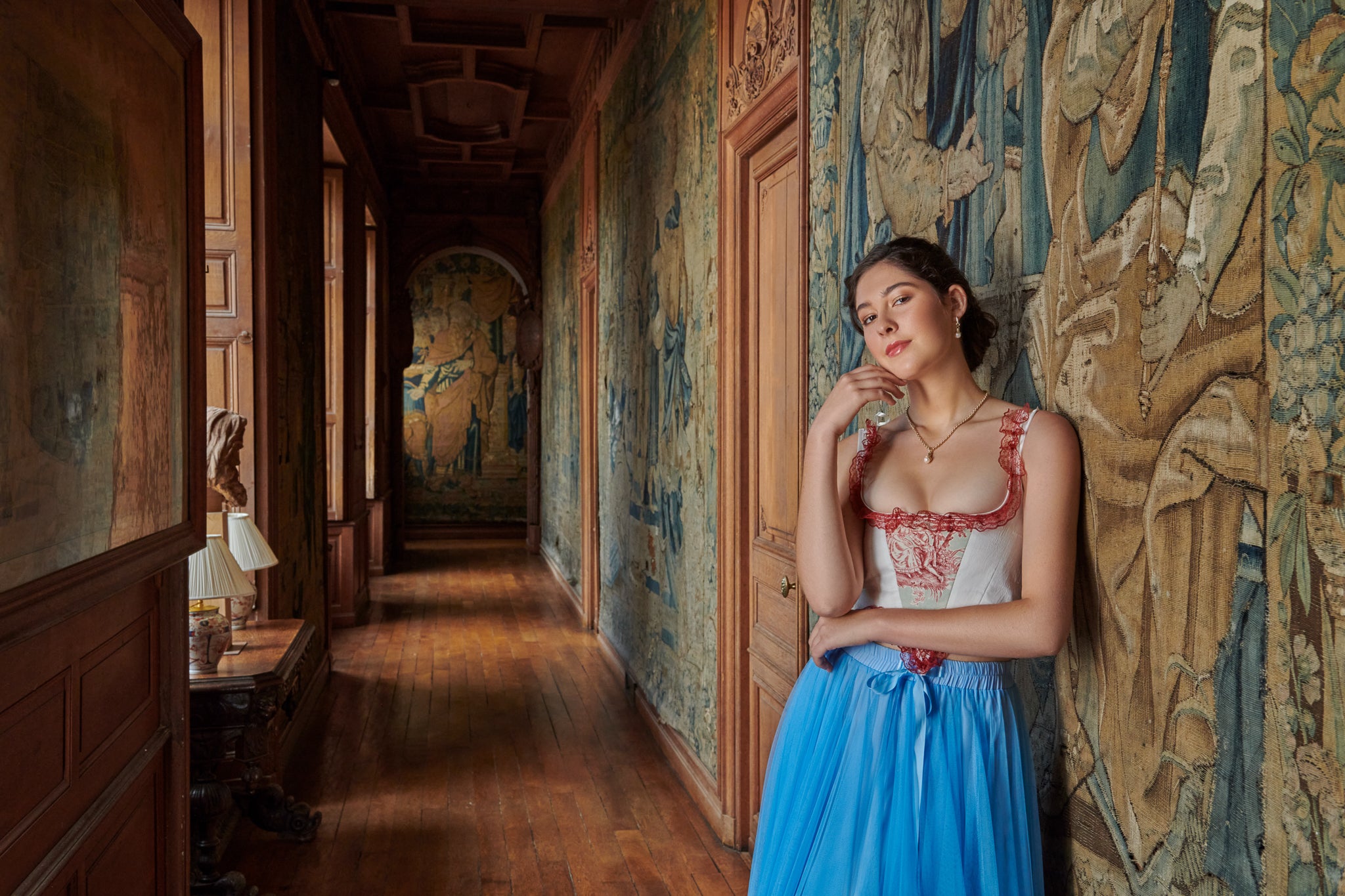 Model wearing handcrafted gold jewellery inside a château and leaning on a tapestry.