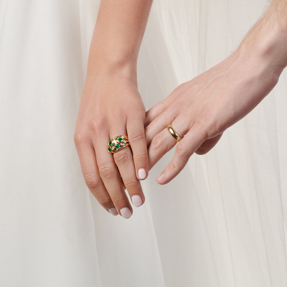 A woman's hand gently touching a man's hand, showcasing a handcrafted 18ct yellow gold engagement ring set with precious stones on hers, alongside a yellow gold wedding band on his.