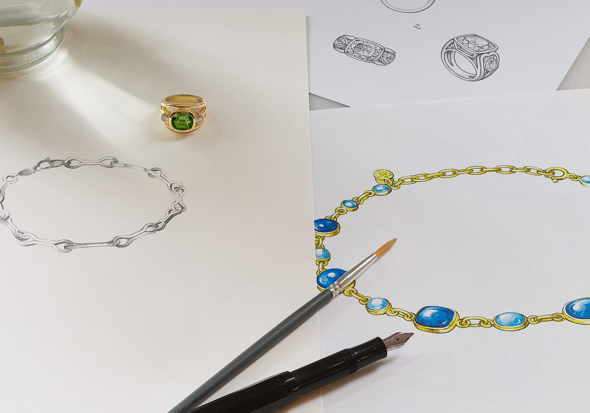 An 18ct yellow gold ring set with diamonds and a green precious stone, placed on top of jewellery sketches featuring necklaces and rings.