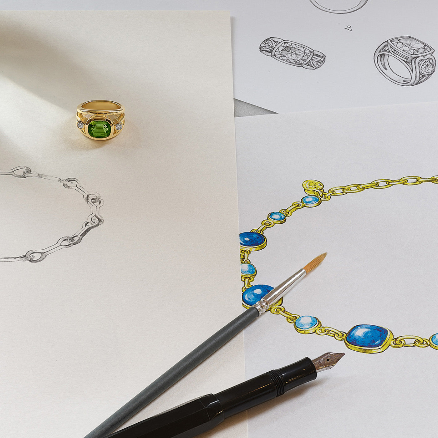 An 18ct yellow gold ring set with diamonds and a green precious stone, placed on top of jewellery sketches featuring necklaces and rings.